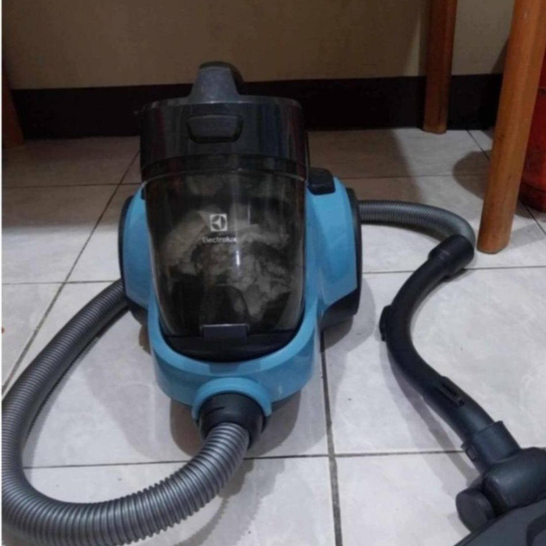 Electrolux Vacuum Repair 1 The image shows a light blue canister vacuum cleaner. The dust cup is visible and appears to be full of dust and debris. The vacuum cleaner is on a tiled floor. A portion of a wooden table leg is visible in the upper right corner.