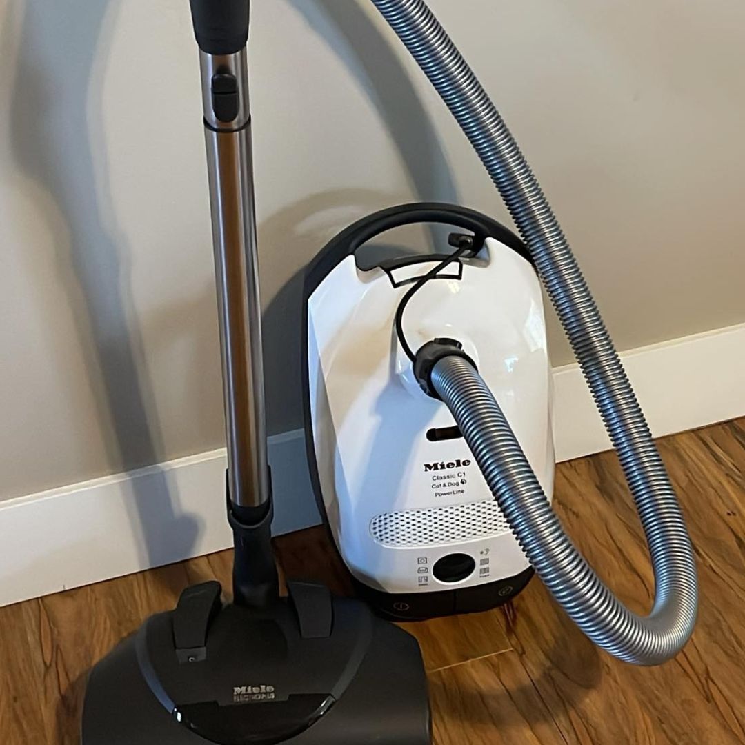 Miele Vacuum Repair 2 The photo shows two Miele vacuum cleaners. A white canister vacuum, a Miele Classic C1 Cat & Dog PowerLine, is positioned next to a black upright vacuum cleaner, also a Miele.