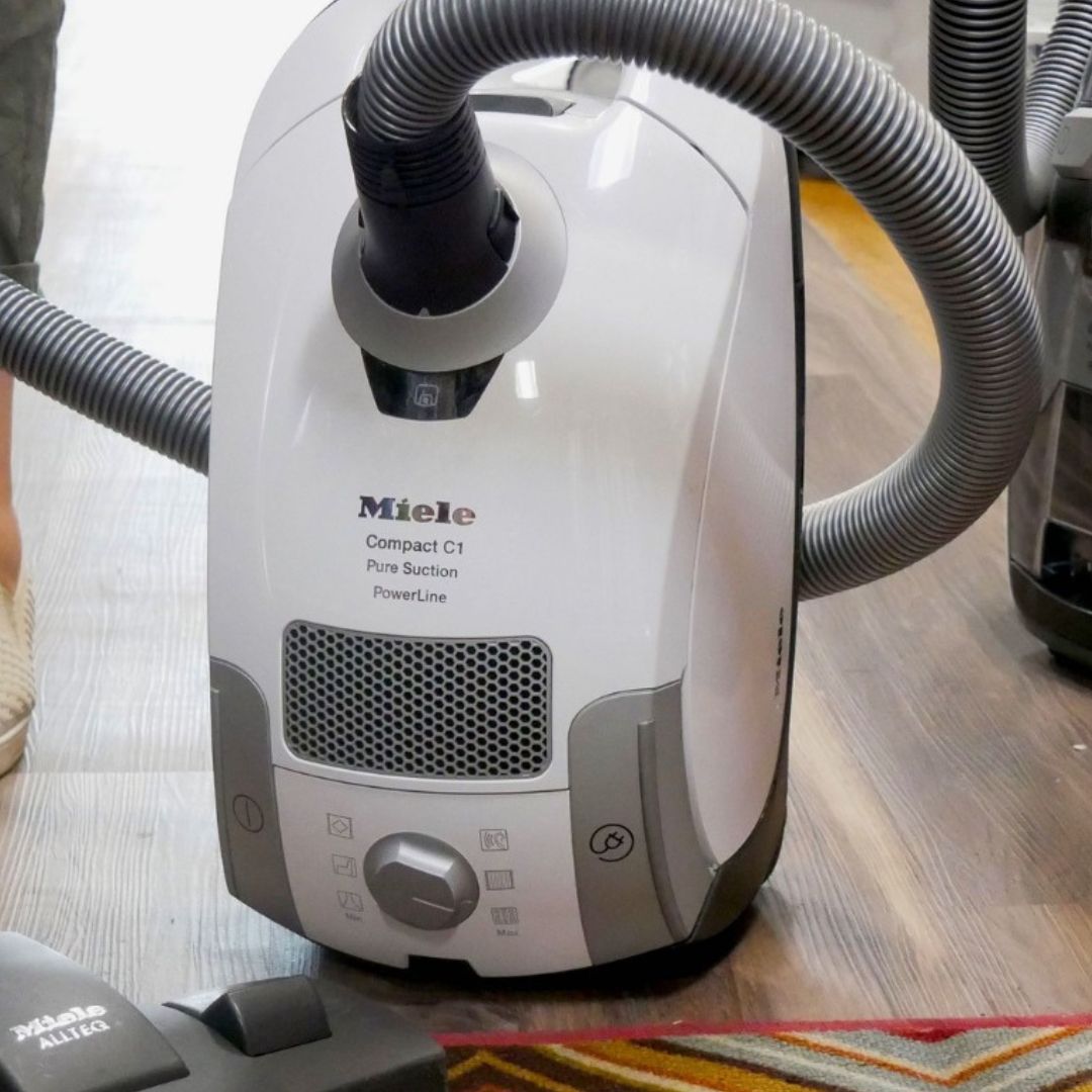 Miele Vacuum Repair a Miele Compact C1 Pure Suction PowerLine canister vacuum cleaner. The image shows the vacuum on a hardwood floor, with its hose partially visible, and a portion of a rug or carpet in the bottom right corner.