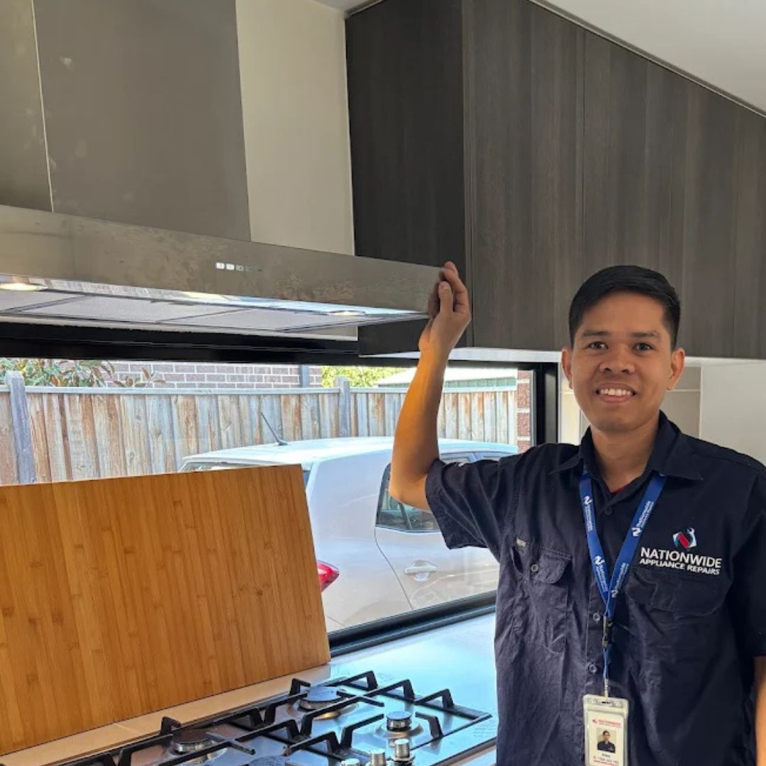 Rangehood Repairs 1 The photo shows a smiling man in a dark blue Nationwide Appliance Repairs uniform. He's standing in a modern kitchen, his arm raised as if he's just installed or is inspecting a stainless steel range hood above a gas cooktop. A bamboo cutting board is partially visible on the counter. A window behind him shows the exterior of a house.