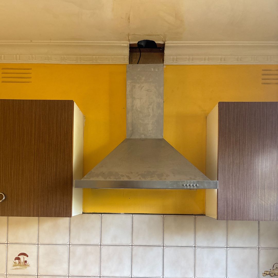Rangehood Repairs 2 The image shows a stainless steel kitchen range hood mounted against a yellow wall. The hood is above a tiled backsplash with a subtle mushroom design. Dark brown wooden cabinets are on either side of the hood. The ceiling shows some minor damage or discoloration around the hood's vent. The overall style appears dated or retro.