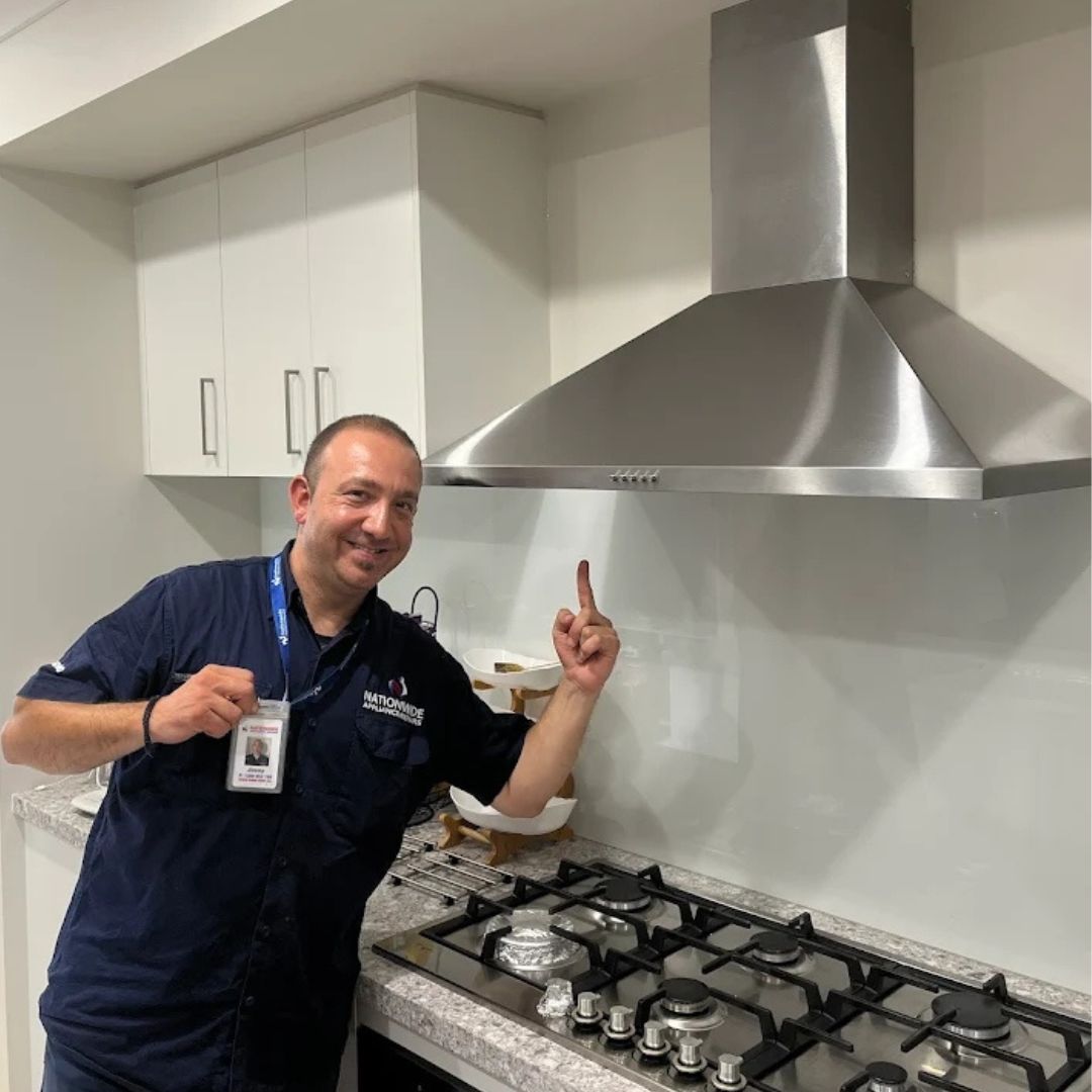 Rangehood Repairs 3 The photo shows a man in a dark blue uniform, pointing upward towards a stainless steel range hood installed above a gas cooktop in a modern kitchen. He's holding an ID badge and appears to be a technician or installer. The kitchen has white cabinets and a light-colored countertop.