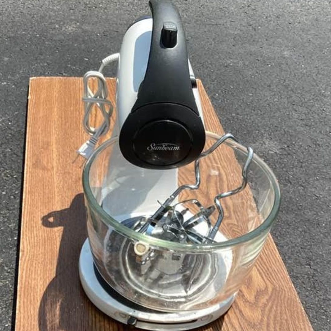 sunbeam-mixer-repair1 Sunbeam stand mixer. It's primarily white with a black handle and base. The mixer's beaters are inside a clear glass bowl, which sits on a white base. The mixer appears to be used, and the photo is taken outdoors on a wooden surface.